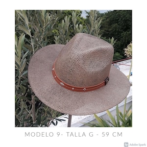 Op de afbeelding: Een geweven strohoed met een bruine leren band versierd met zilverkleurige studs. De hoed heeft een brede rand en een klassieke fedora-vorm. De hoedmaat is 59 cm.