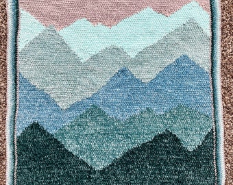 May include: A crocheted wall hanging featuring a mountain range with a pink, blue, and green color scheme.