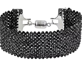 May include: A wide bracelet composed of multiple rows of small, faceted black beads arranged in a woven pattern. The bracelet features a polished silver-tone metal clasp with a cylindrical magnetic closure mechanism at the center of the design.