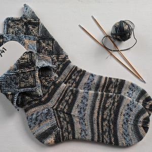 Könnte beinhalten: Handgestrickte Socken in Blau-, Grau- und Beigetönen mit geometrischem Muster. Ein weißes Etikett mit der Größe 40/41 ist in die Socke gesteckt. Stricknadeln und ein Knäuel Wolle liegen neben den Socken.