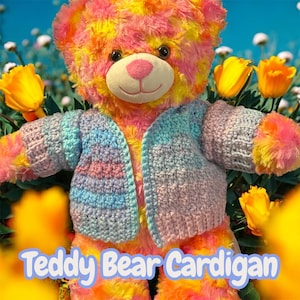 May include: A multi-coloured teddy bear wearing a crocheted cardigan. The bear has a pink nose and is surrounded by yellow flowers. The cardigan is a mix of blue, pink, and grey. The text "Teddy Bear Cardigan" is at the bottom.