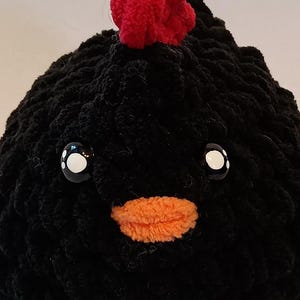May include: Black crocheted chicken plush toy with orange beak and white eyes. The chicken has a red crocheted comb on top of its head.