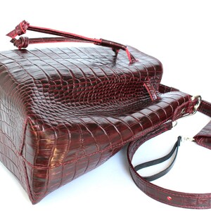 May include: A burgundy leather tote bag with a crocodile embossed pattern. The bag has a top handle and a long adjustable strap. It also has a small, detachable pouch with a snap closure.
