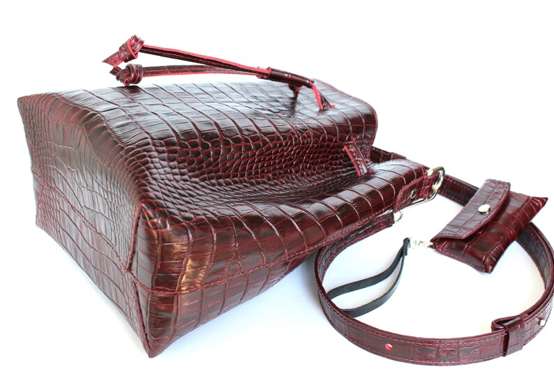 May include: A burgundy leather tote bag with a crocodile embossed pattern. The bag has a top handle and a long adjustable strap. It also has a small, detachable pouch with a snap closure.