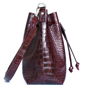 May include: A burgundy leather bucket bag with a crocodile embossed pattern. The bag has a long adjustable strap and a drawstring closure.