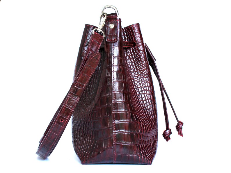 May include: A burgundy leather bucket bag with a crocodile embossed pattern. The bag has a long adjustable strap and a drawstring closure.