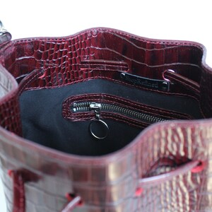 May include: A burgundy leather bucket bag with a crocodile embossed pattern. The bag has a drawstring closure and a zippered pocket inside. The bag is lined with black fabric and has a silver metal ring attached to the zipper.