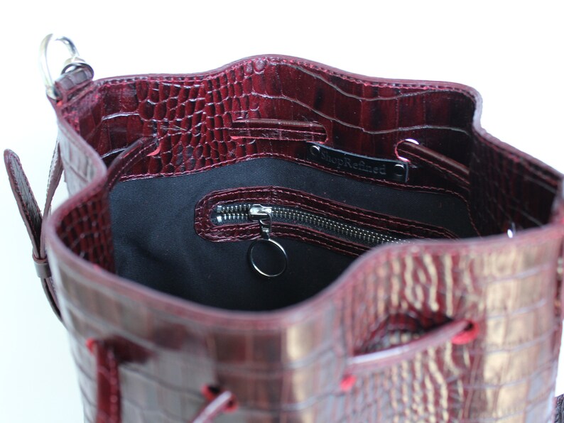 May include: A burgundy leather bucket bag with a crocodile embossed pattern. The bag has a drawstring closure and a zippered pocket inside. The bag is lined with black fabric and has a silver metal ring attached to the zipper.