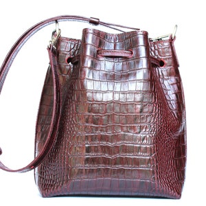 May include: A burgundy leather bucket bag with a crocodile embossed pattern and a long adjustable strap.