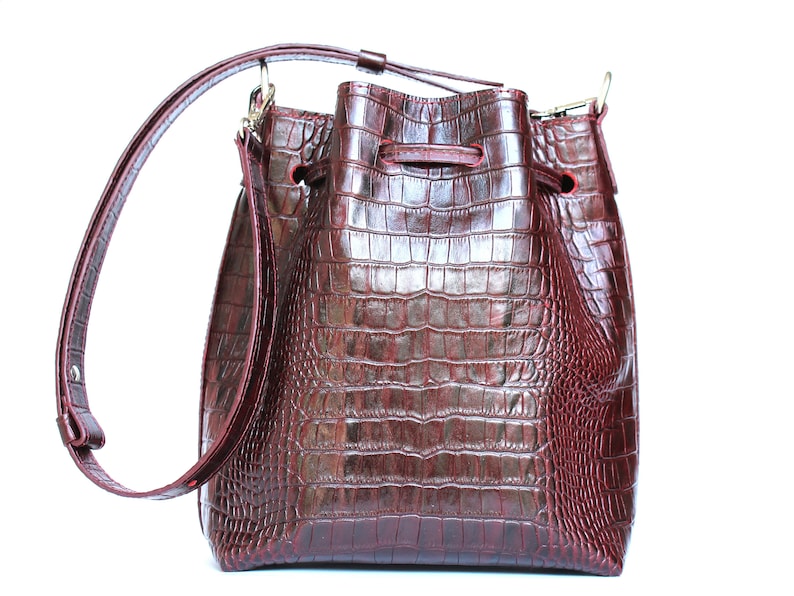 May include: A burgundy leather bucket bag with a crocodile embossed pattern and a long adjustable strap.