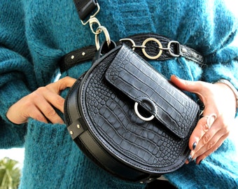 May include: A black crocodile-embossed leather satchel bag with a silver buckle and a silver chain strap. The bag is worn over a teal blue fuzzy sweater.