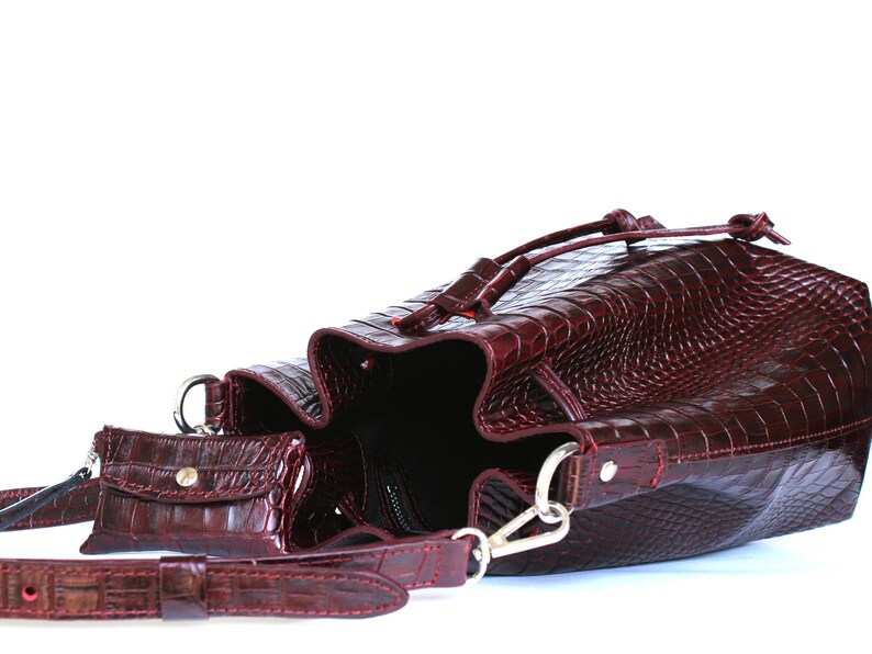 May include: A burgundy leather bucket bag with a drawstring closure and a matching small pouch. The bag has a crocodile-embossed pattern.