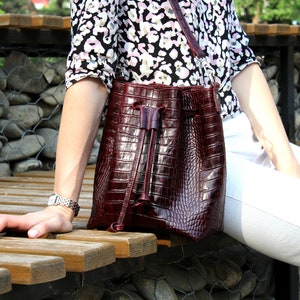 May include: A burgundy leather bucket bag with a drawstring closure. The bag has a crocodile-embossed texture.