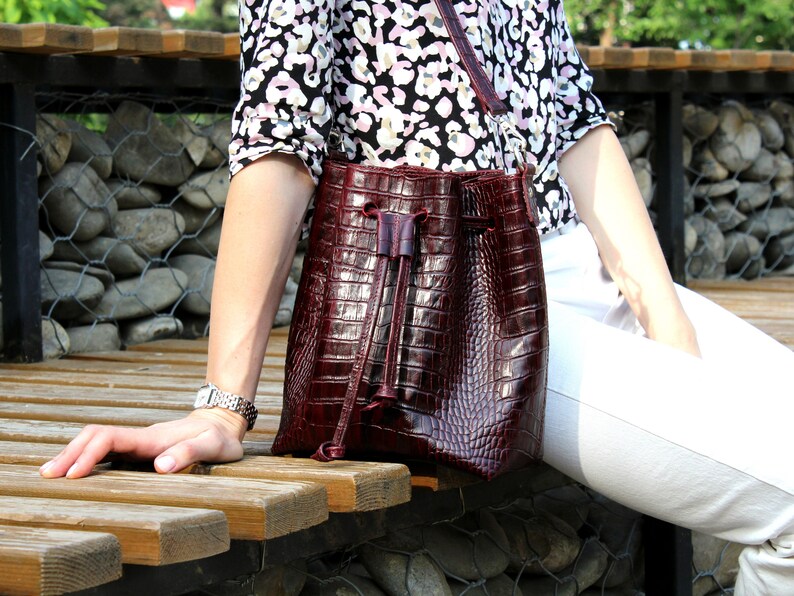 May include: A burgundy leather bucket bag with a drawstring closure. The bag has a crocodile-embossed texture.
