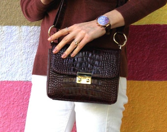 May include: A brown crocodile-embossed leather crossbody bag with a gold clasp and a gold chain strap. The bag is worn over a white shirt and brown sweater.