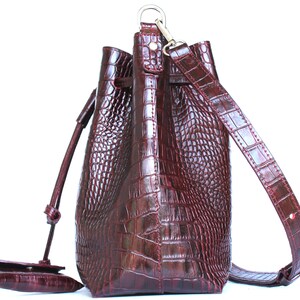 May include: A burgundy leather bucket bag with a long adjustable strap and a drawstring closure. The bag has a crocodile embossed pattern.