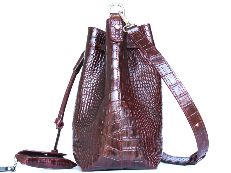 May include: A burgundy leather bucket bag with a long adjustable strap and a drawstring closure. The bag has a crocodile embossed pattern.