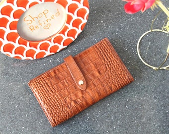 May include: A brown leather wallet with a crocodile embossed pattern and a gold snap closure. The wallet has a flap with a strap and a gold buckle.