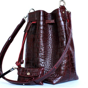May include: A burgundy leather bucket bag with a crocodile embossed pattern and a long adjustable strap. The bag has a drawstring closure and a small matching pouch.