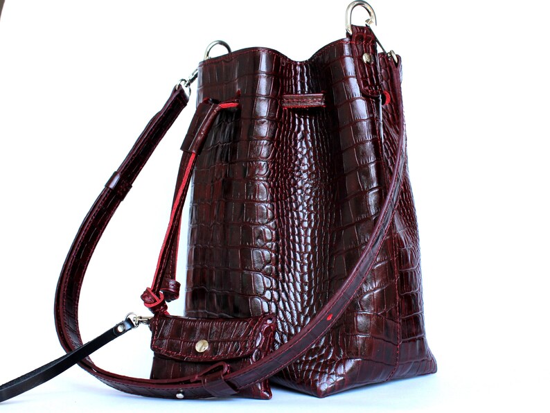May include: A burgundy leather bucket bag with a crocodile embossed pattern and a long adjustable strap. The bag has a drawstring closure and a small matching pouch.