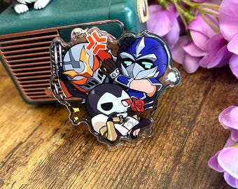 May include: A clear acrylic charm featuring three stylized cartoon characters. One character wears a blue and silver mask, another has an orange and black helmet with an angry symbol, and the third has a white skull mask and holds a red rose.