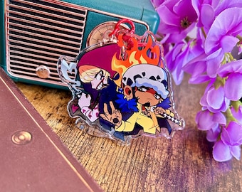 May include: A clear acrylic keychain featuring a colorful anime-style illustration of two characters. One character wears a red hood, while the other has a white spotted hat, yellow shirt, and holds a sword. The design includes orange flame accents.
