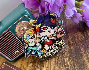May include: A clear acrylic charm featuring a colorful anime-style illustration of two characters with dark and blonde hair, red eyes, and decorative headpieces, surrounded by small skull motifs and a gold metal jump ring attachment.