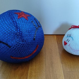 May include: Two whimsical, round fabric dolls. One is a large blue sequined ball with a stitched face and a red star. The other is a smaller white ball with a stitched face and a red bow.