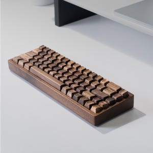 Puede incluir: Un teclado de madera con teclas marrones oscuras y un marco de madera a juego. Las teclas están dispuestas en un diseño estándar. El teclado está diseñado para una estética minimalista y se encuentra sobre una superficie blanca.