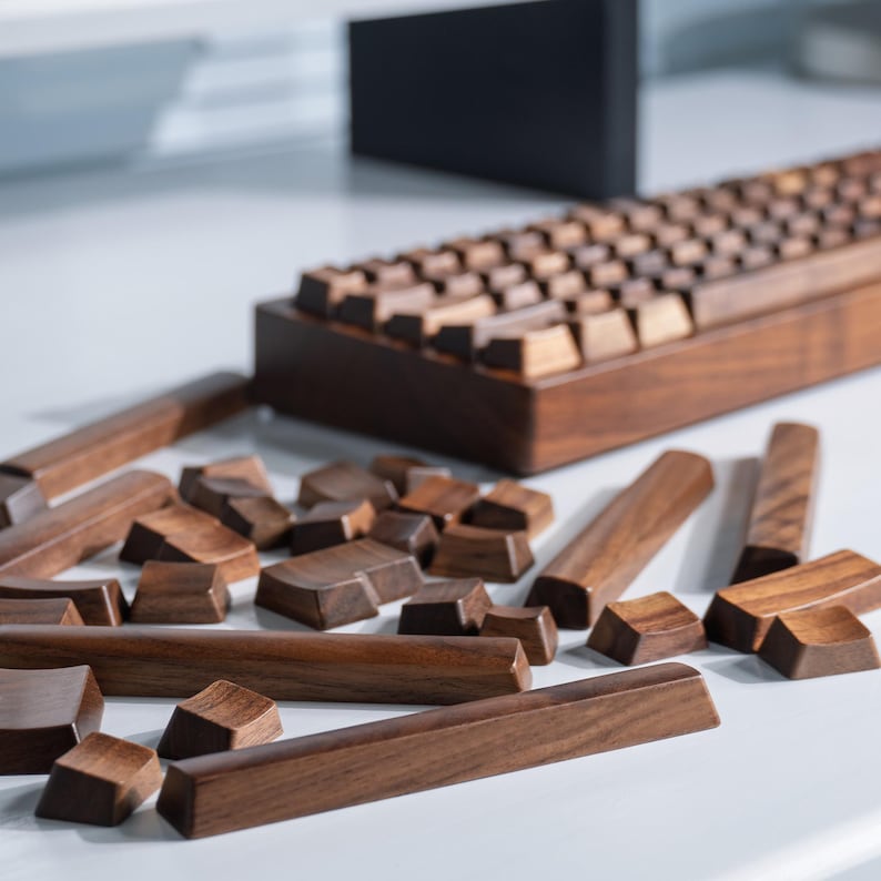 May include: A wooden keyboard with keycaps and individual keys scattered in front. The keyboard and keys are made of dark brown wood. The keyboard has a rectangular shape with a slightly angled design.