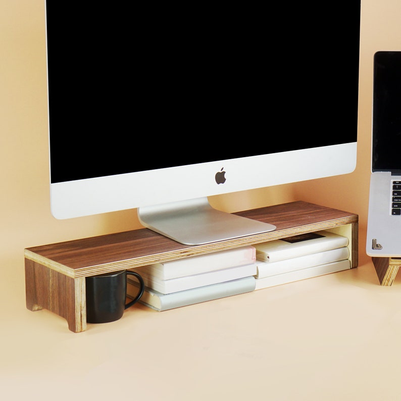 May include: A wooden monitor stand with a dark brown finish. The stand is raised on two legs and has a shelf for storing books or other items. A computer monitor and a laptop are sitting on the stand.