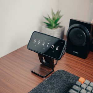 An iPhone is placed horizontally on the 3 in 1 fold charger, with a digital clock displayed on the screen, surrounded by potted plants, stereo, keyboard, etc., placed on the walnut table is very beautiful.