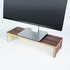 May include: A wooden monitor stand with a dark brown top and light brown sides. A silver computer monitor is sitting on the stand.