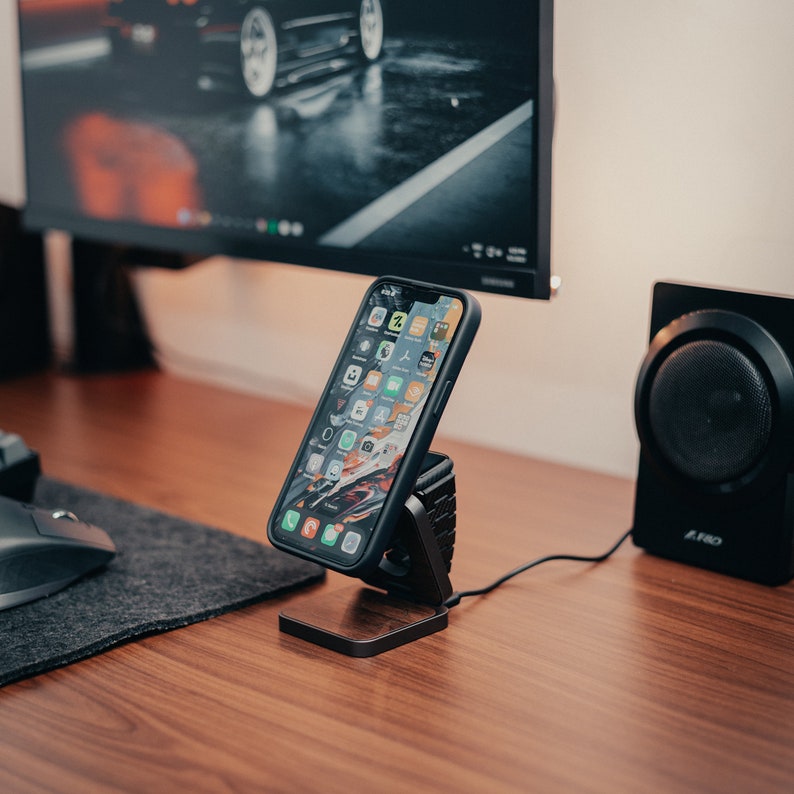 An iPhone and Apple watch sits on a 3-in-1 charger on a walnut surface, surrounded by a computer, keyboard, and impressions, which look pretty on a walnut-colored desktop.