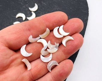 May include: A collection of white mother of pearl crescent moon shaped charms. The charms are scattered on a hand and a dark gray surface.