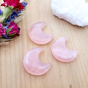 May include: Three pink crescent moon shaped rose quartz crystals on a wooden surface.