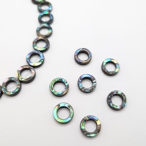 May include: A strand of iridescent abalone shell beads in a circle shape. The beads are strung on a clear thread and are scattered on a white surface.