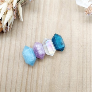 May include: Four faceted gemstone beads in shades of blue, purple, white, and green. The beads are arranged in a row on a wooden surface.