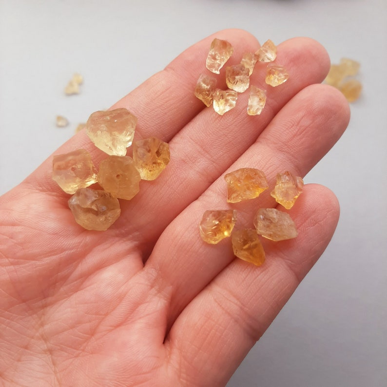 Pu&ograve; includere: Una collezione di piccoli cristalli di citrino grezzi, di colore giallo-arancione. I cristalli sono di varie forme e dimensioni e sono mostrati su una mano.