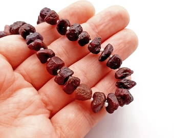 Puede incluir: Una hebra de cuentas de piedras preciosas de color rojo oscuro, probablemente rubíes, enhebradas en un hilo blanco. Las cuentas tienen una forma irregular y una textura rugosa y natural.