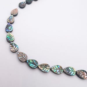 May include: A necklace of iridescent leaf-shaped beads. The beads have a pearlescent sheen with green, blue, and brown hues. Each bead is carved with leaf veins. The beads are strung on a clear string, creating a delicate and natural appearance.
