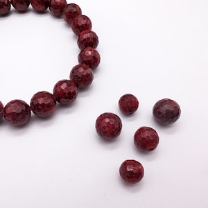 May include: A strand of dark red gemstone beads, with a few loose beads scattered around it. The beads are faceted and have a rough, textured surface.