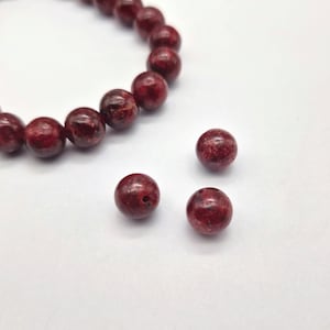 May include: A bracelet and loose beads are shown in a close-up. The beads are a deep red colour with dark speckles. The bracelet is strung with the same beads. The beads are round and have a matte finish.