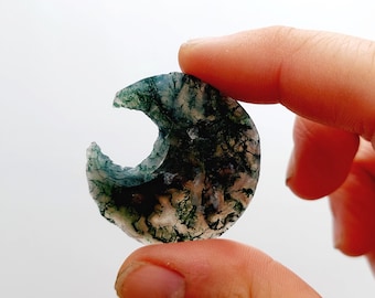 May include: A crescent-shaped green agate stone with a white and green pattern. The stone is translucent and has a natural, rough texture.