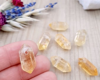 May include: Seven small, clear, yellow-gold, faceted, crystal points with a drilled hole at the top. The points are arranged on a light wood surface.