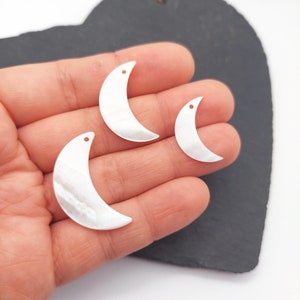 May include: Three white mother of pearl crescent moon charms with a hole for stringing. The charms are different sizes and are shown in a hand.