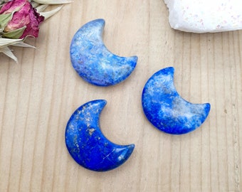 May include: Three blue crescent moon shaped lapis lazuli crystal stones on a wooden surface.
