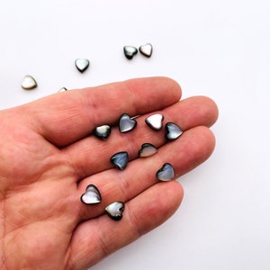 May include: A collection of small, iridescent black and white heart-shaped shell beads. The beads are scattered on a hand and a white surface.