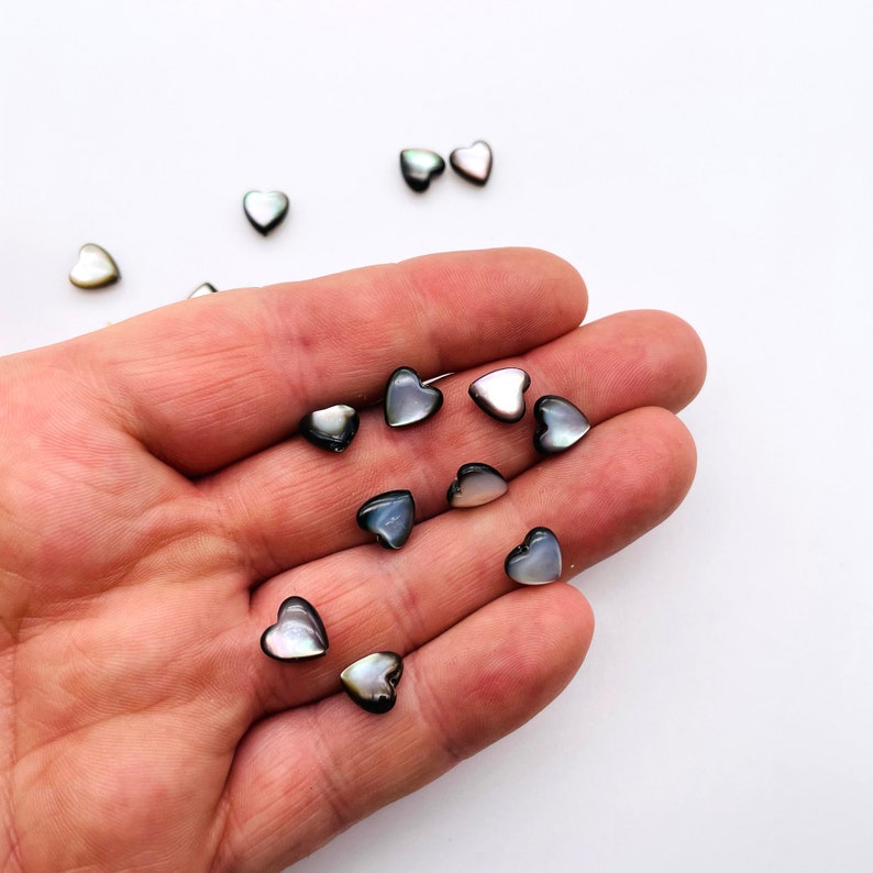 May include: A collection of small, iridescent black and white heart-shaped shell beads. The beads are scattered on a hand and a white surface.