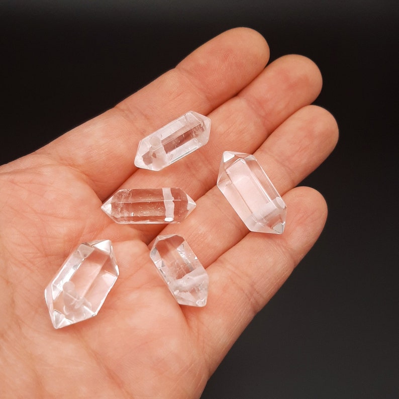 May include: Five clear quartz crystal points in the shape of double-terminated wands, displayed on a hand.